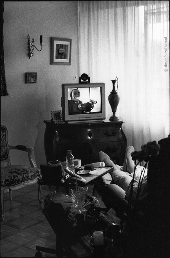 Photographie noir et blanc d'un salon d'appartement avec meuble télévision et fauteuil. Sur l'écran apparait l'image d'un homme masqué tenant un pistolet dans la direction du spectateur assis dans son fauteuil.