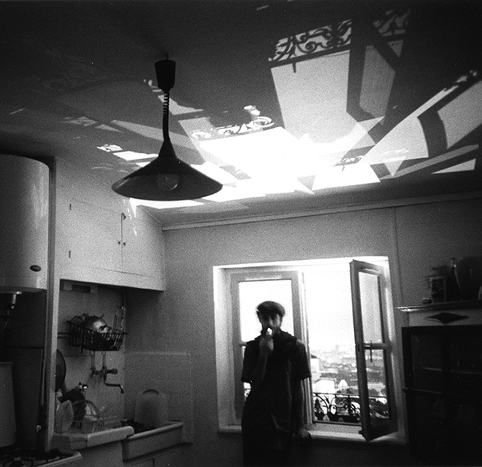 Photographie noir et blanc montrant Simon Bezzi Batani debout devant un fenêtre dans une pièce. Une forte lumière entre par la fenêtre et projette sur le plafond, de façon très graphique et florale, des ombres de ferronnerie et d'encadrement de fenêtre.